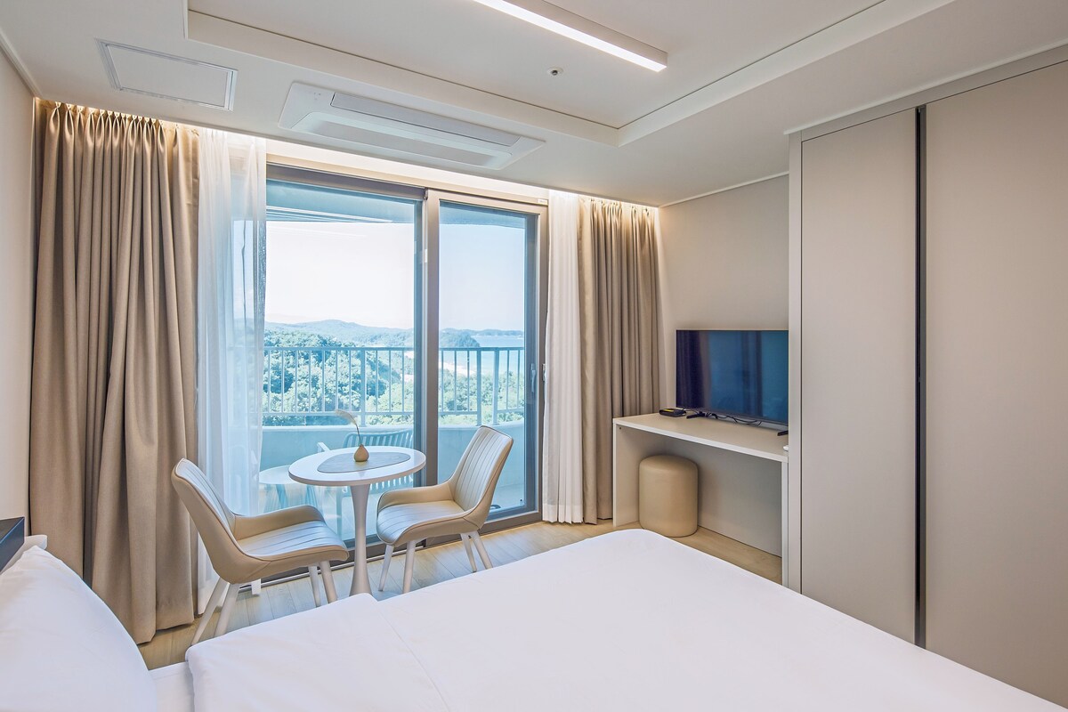 A hotel room with a bed, a TV, a small table with chairs, a balcony with curtains, and a view of the city.