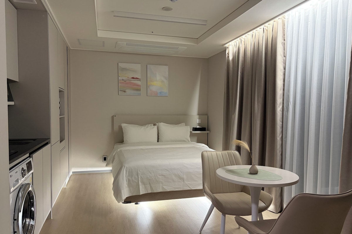 Habitación de hotel con cama, una mesita, una silla, lavadora y ventana con cortinas.