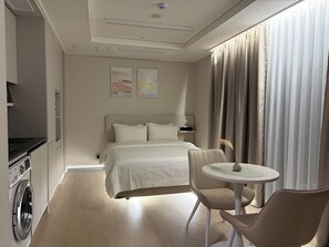 Habitación de hotel con cama, una mesita, una silla, lavadora y ventana con cortinas.