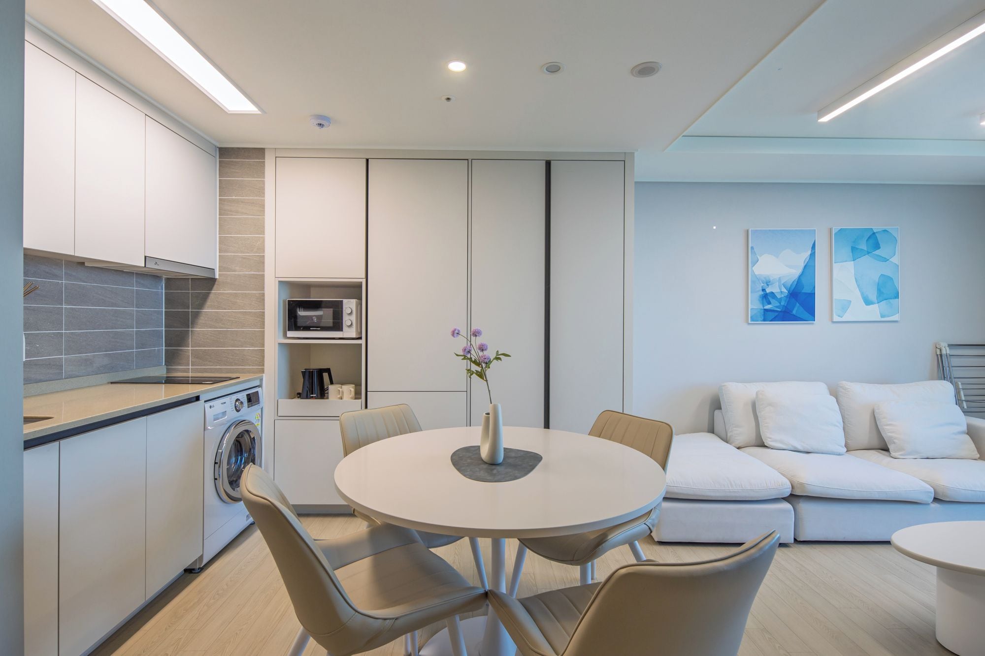 A modern kitchen with built-in appliances, a dining area with a round table and chairs, and a living area with a sofa and abstract wall art.