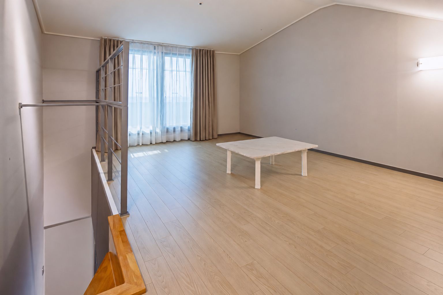 A spacious room with a wooden floor, a white table, and a staircase with a metal railing.