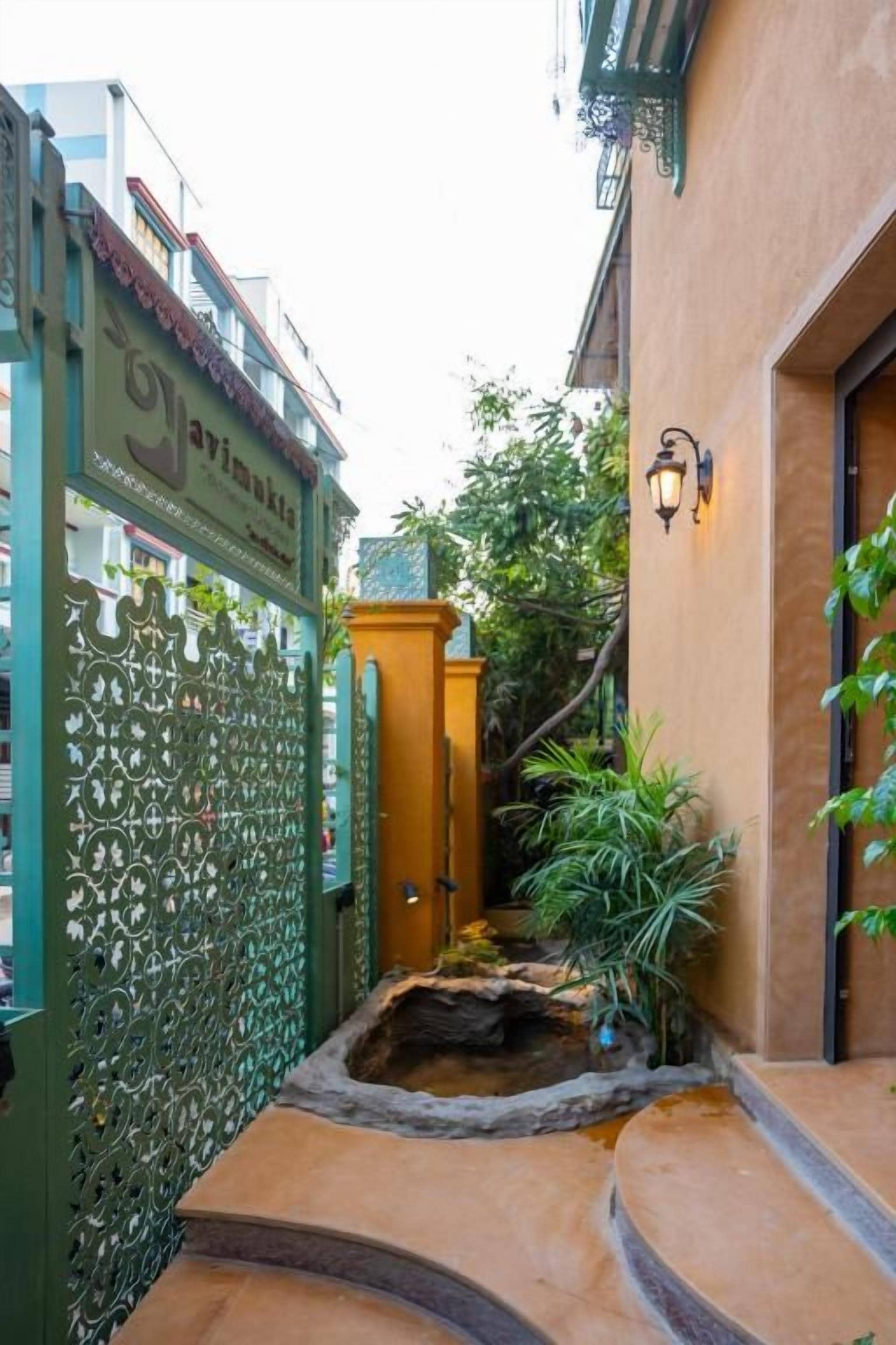 A narrow alleyway with a green gate, a small water feature, and a building entrance with a light fixture.