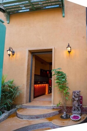 Entrada de un edificio con una escalera de piedra, un pequeño jardín con una estatua y dos luces fijadas a la pared.