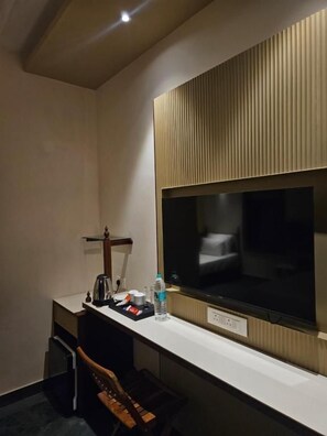 Habitación de hotel con televisor de pantalla plana grande montado en la pared, un escritorio pequeño con hervidor eléctrico, una botella de agua y una silla.