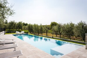 A rectangular swimming pool with clear blue water, surrounded by a low stone wall. There are white lounge chairs and a small table on the poolside. - Apartment Groppello Casa Franzosiwith pool (Puegnago sul Garda)