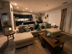 Ein modernes Wohnzimmer mit einem beigefarbenen Ecksofa, einem Couchtisch und einem Essbereich, im Hintergrund ist eine Küche zu sehen.