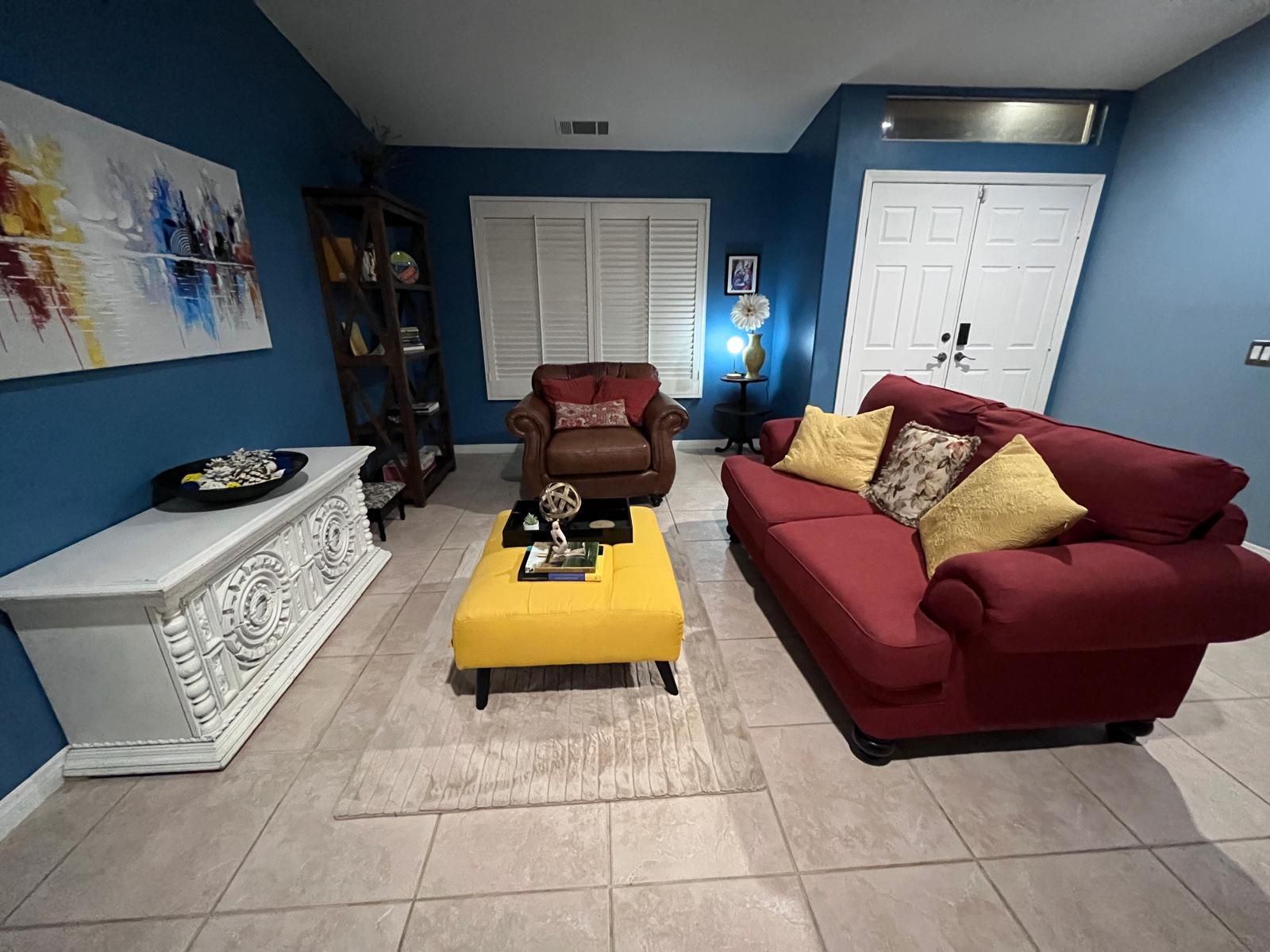 A living room with a red sofa, a yellow ottoman, a brown armchair, a white console table, and a bookshelf.