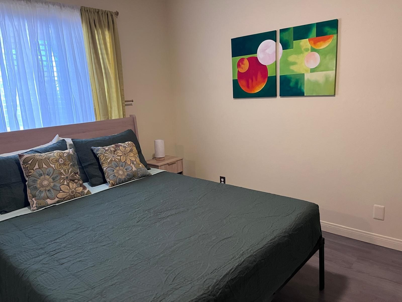 A bedroom with a bed, pillows, a nightstand, a lamp, and two abstract paintings on the wall.