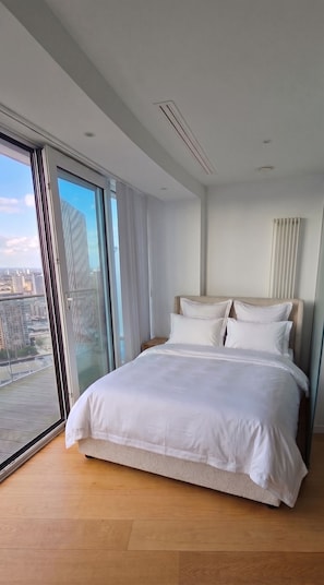A modern bedroom with a large bed, a nightstand, and a view of a cityscape through floor-to-ceiling windows. - Luxury Canary Wharf Skyline Apartment (London)