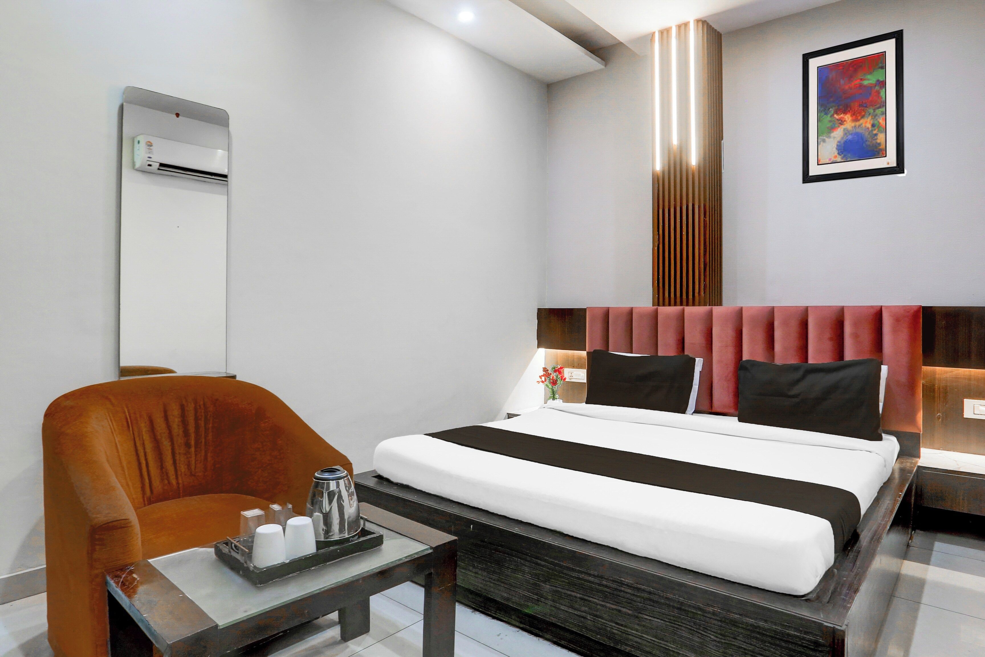 Quarto de hotel com cama, uma cadeira, uma mesa com bandeja de copos, ar condicionado fixado na parede e uma pintura.