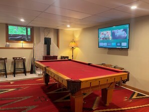 A pool table with a red felt surface, a television mounted on the wall displaying a show, and a window with a view of greenery. - Tranquil 3-bedroom house with AC in charming Conway (Conway)