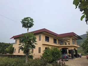 A two-story house with a balcony, surrounded by greenery and parked cars.