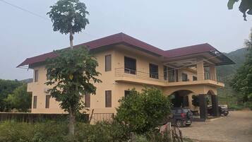 A two-story house with a balcony, a dirt driveway, and a tree in the foreground.