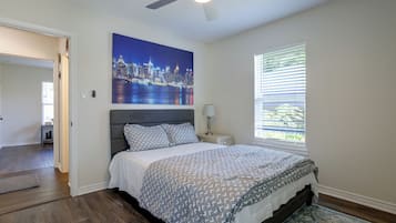 Une chambre à coucher avec un tableau représentant un paysage urbain, un lit avec une literie à motifs, un ventilateur de plafond et une fenêtre avec des stores.