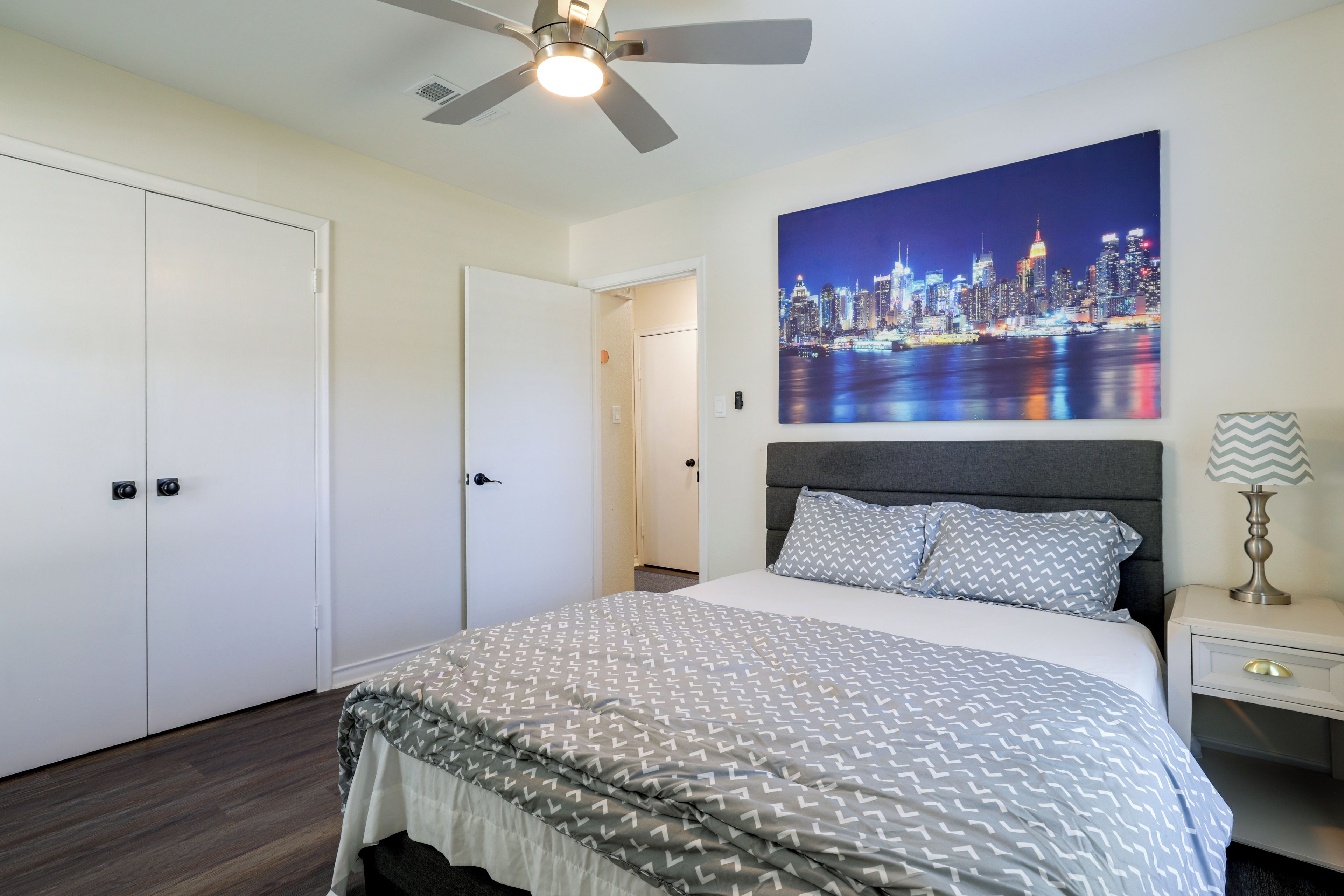 A bedroom with a bed, a ceiling fan, a nightstand, a lamp, and a cityscape painting on the wall.