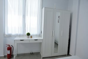 Ein Zimmer mit einem weißen Schreibtisch, einem weißen Kleiderschrank, einem Fenster mit durchsichtigen Vorhängen, einer kleinen Topfpflanze und einem Feuerlöscher.