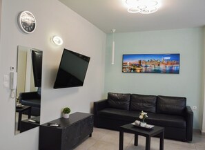 A modern living room with a flat-screen TV, a black sofa, a coffee table, and a cityscape painting on the wall. - Comfortable Central Studios with City Views in Thessaloniki (Thessaloniki)