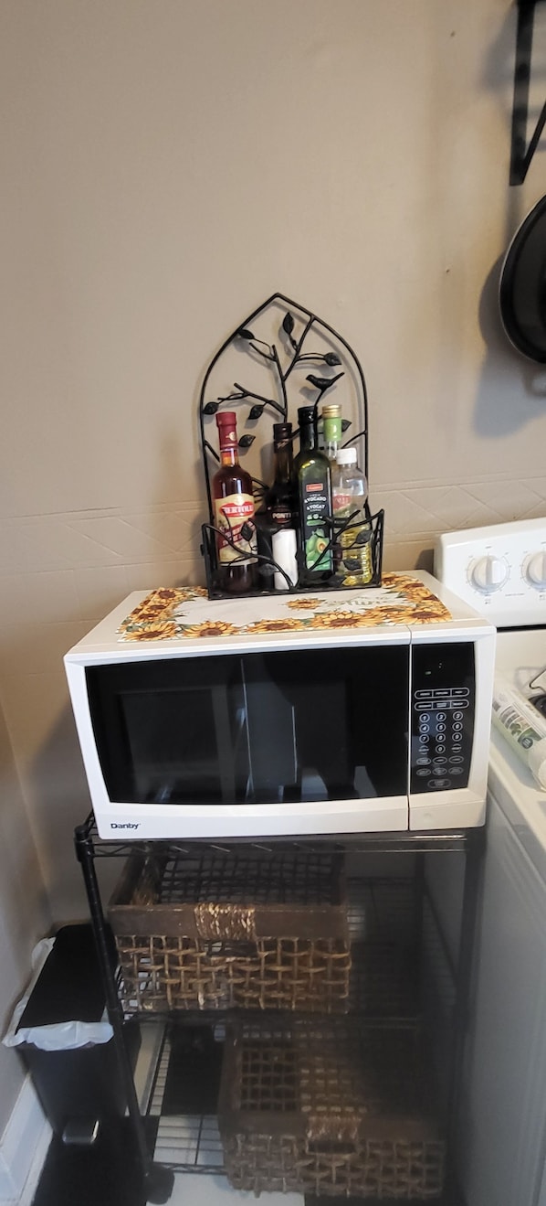 Un four à micro-ondes blanc, surmonté d’une nappe à motifs floraux, une étagère en métal noir avec des bouteilles, et, au fond, une cuisinière blanche.