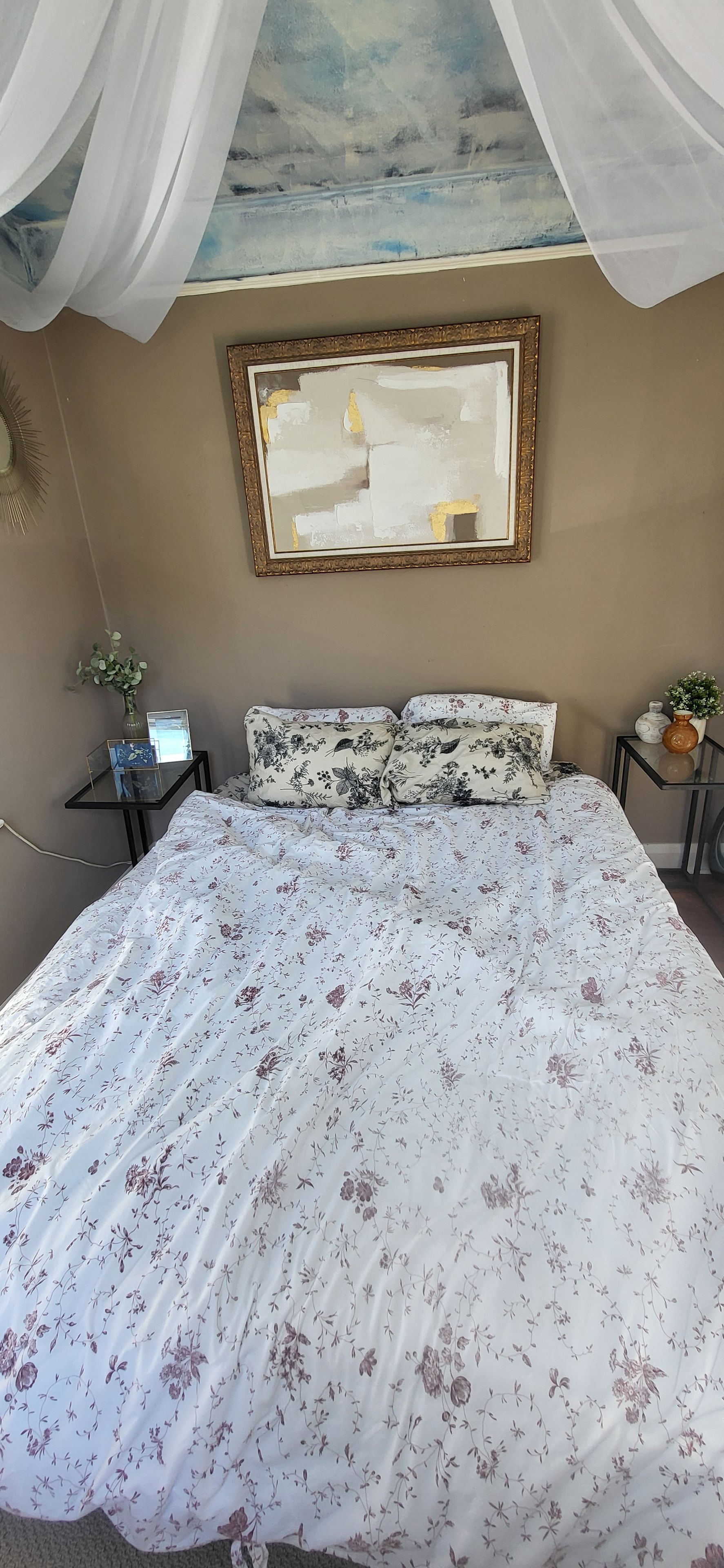 A bedroom with a canopy bed, a framed painting on the wall, and a bedside table with a vase.