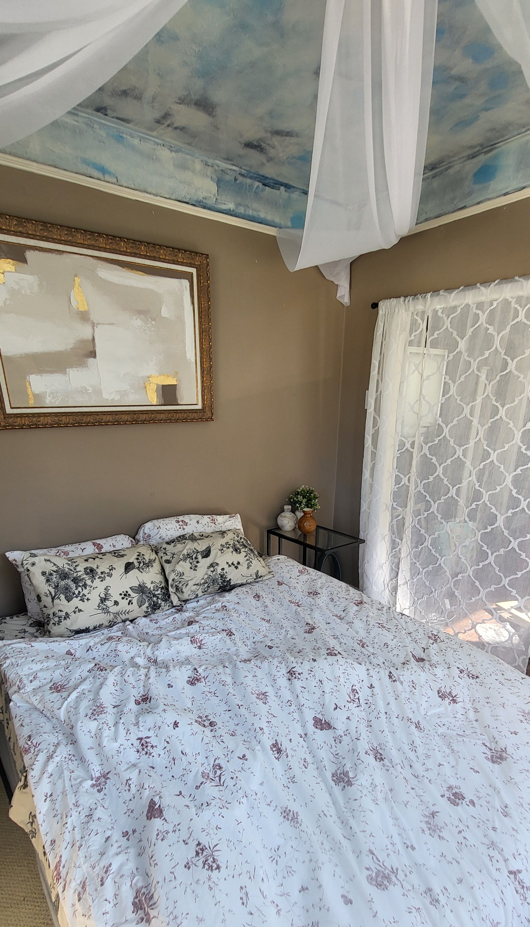 A bedroom with a canopy bed, a framed abstract painting, and a vase with flowers.