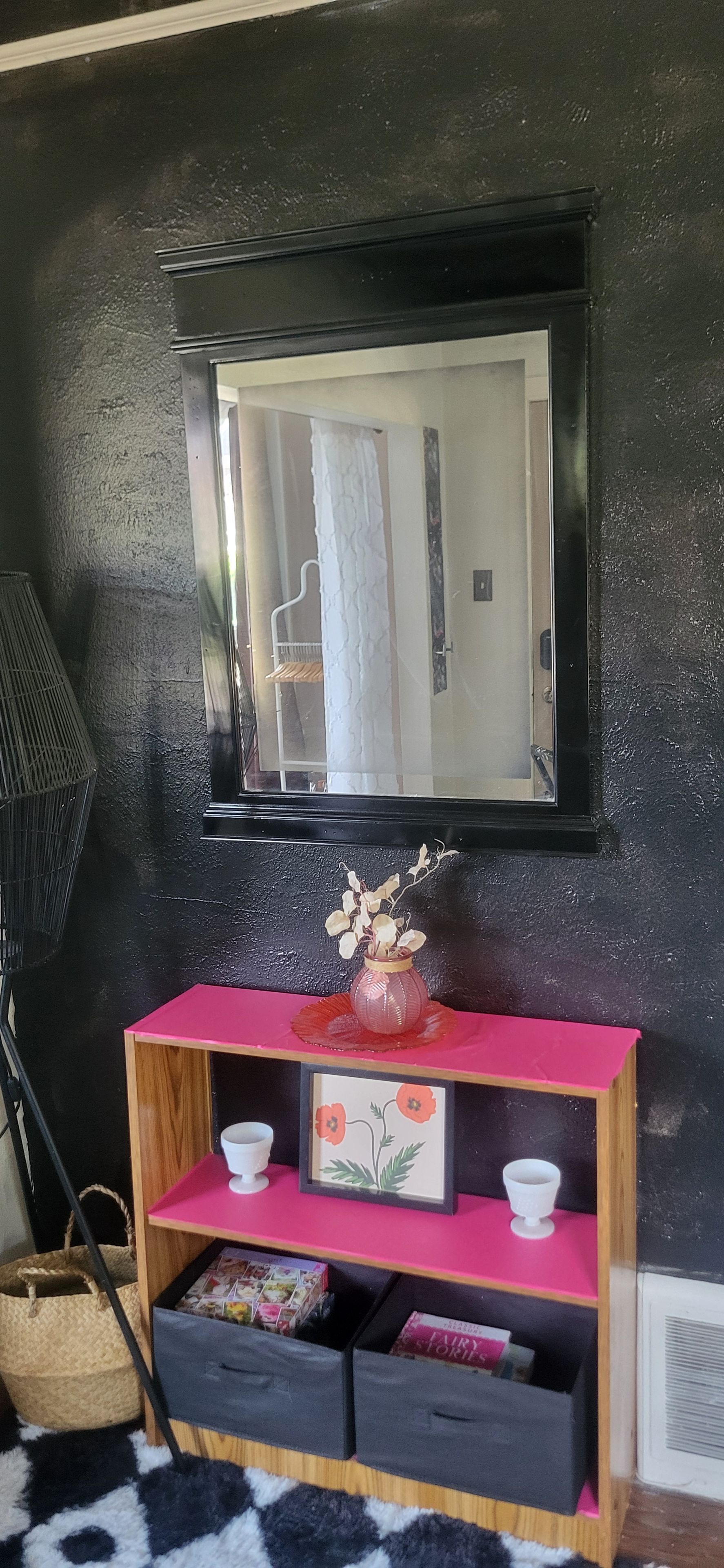 A room with a black wall, a wooden console table with pink top, two white cups, a framed picture, and a small vase with dried flowers.
