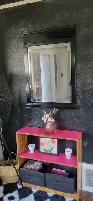 A room with a black wall, a wooden console table with pink top, two white cups, a framed picture, and a small vase with dried flowers.