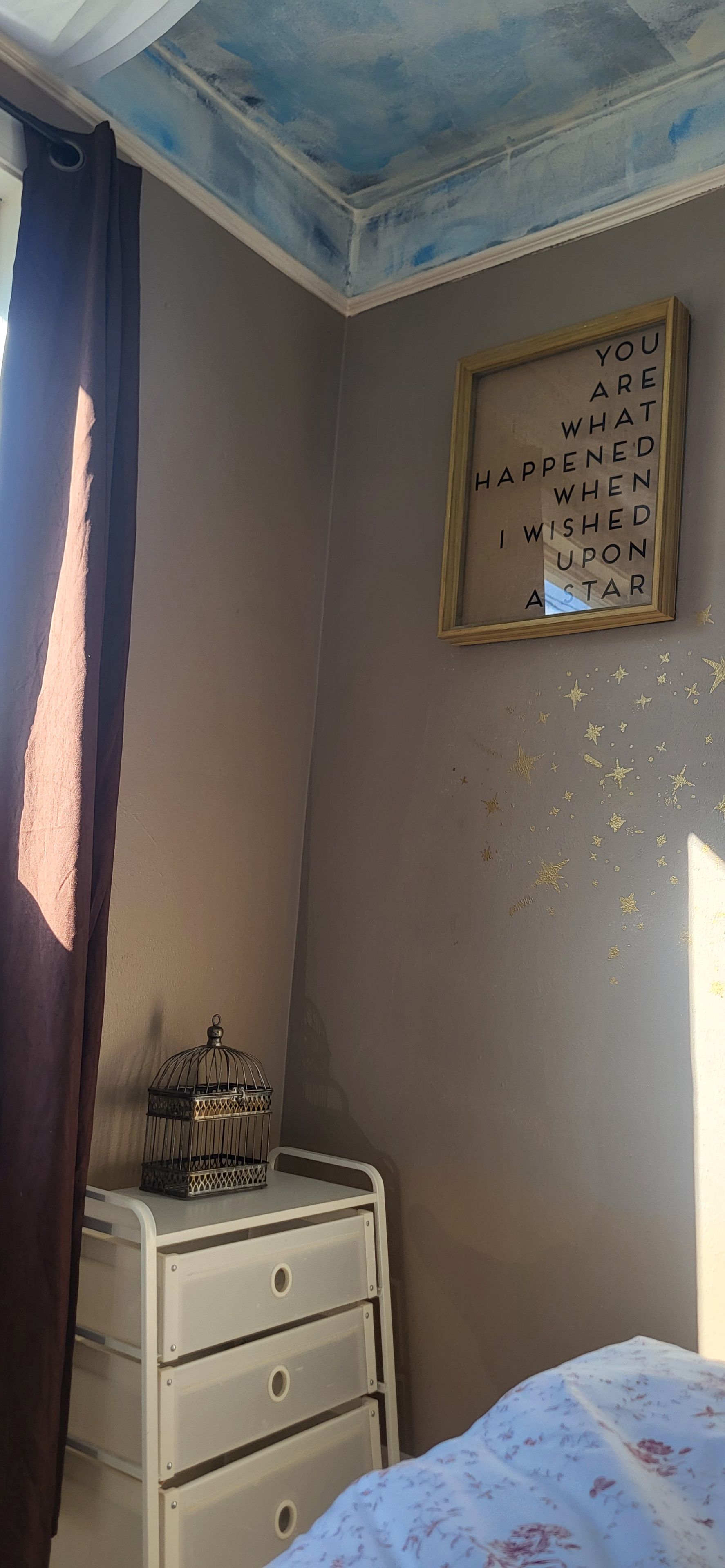 A bedroom with a bed, a nightstand with drawers, a framed inspirational quote on the wall, and a window with a curtain.