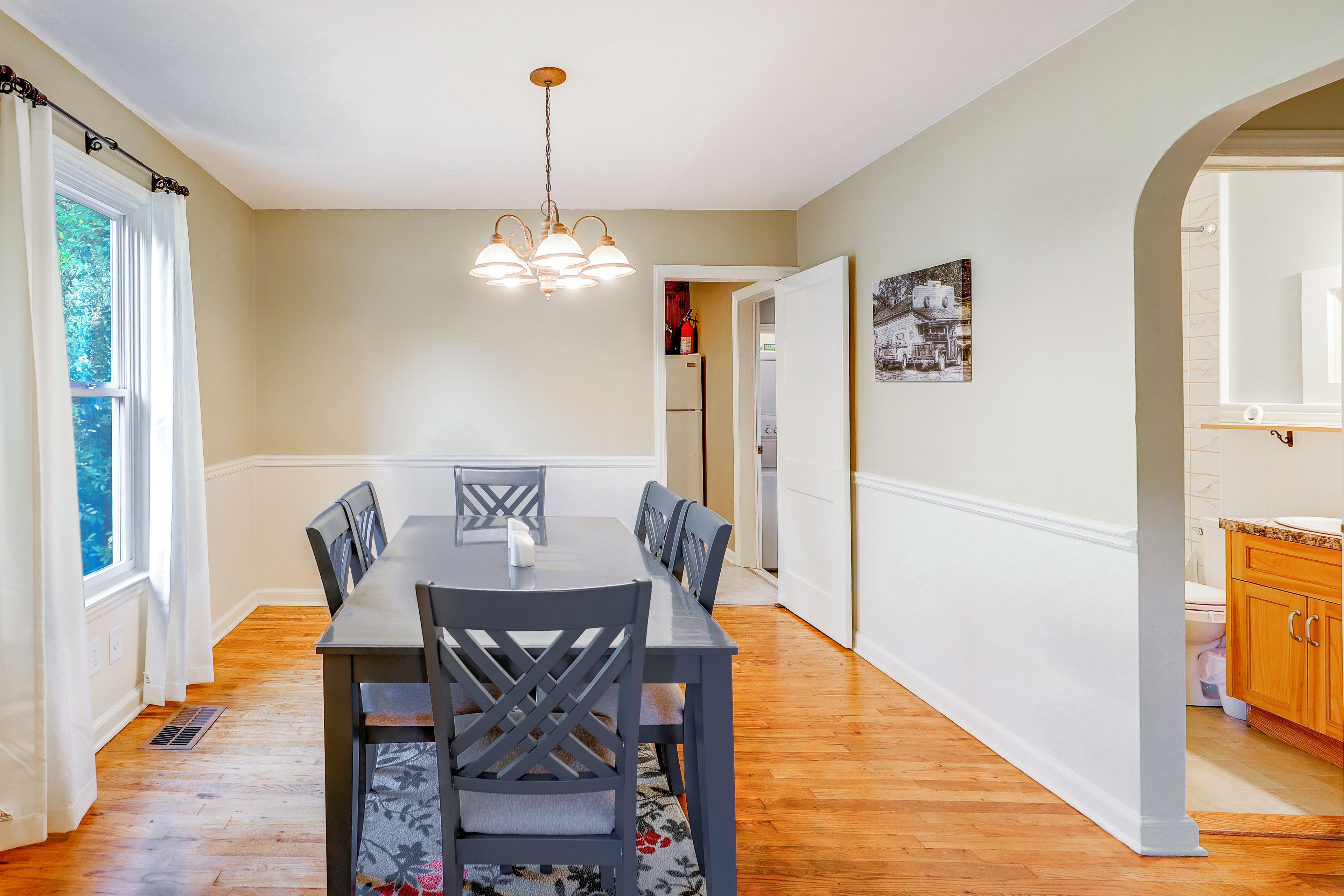 A dining room with a wooden table and chairs, a chandelier, and a view of the kitchen through an open door.