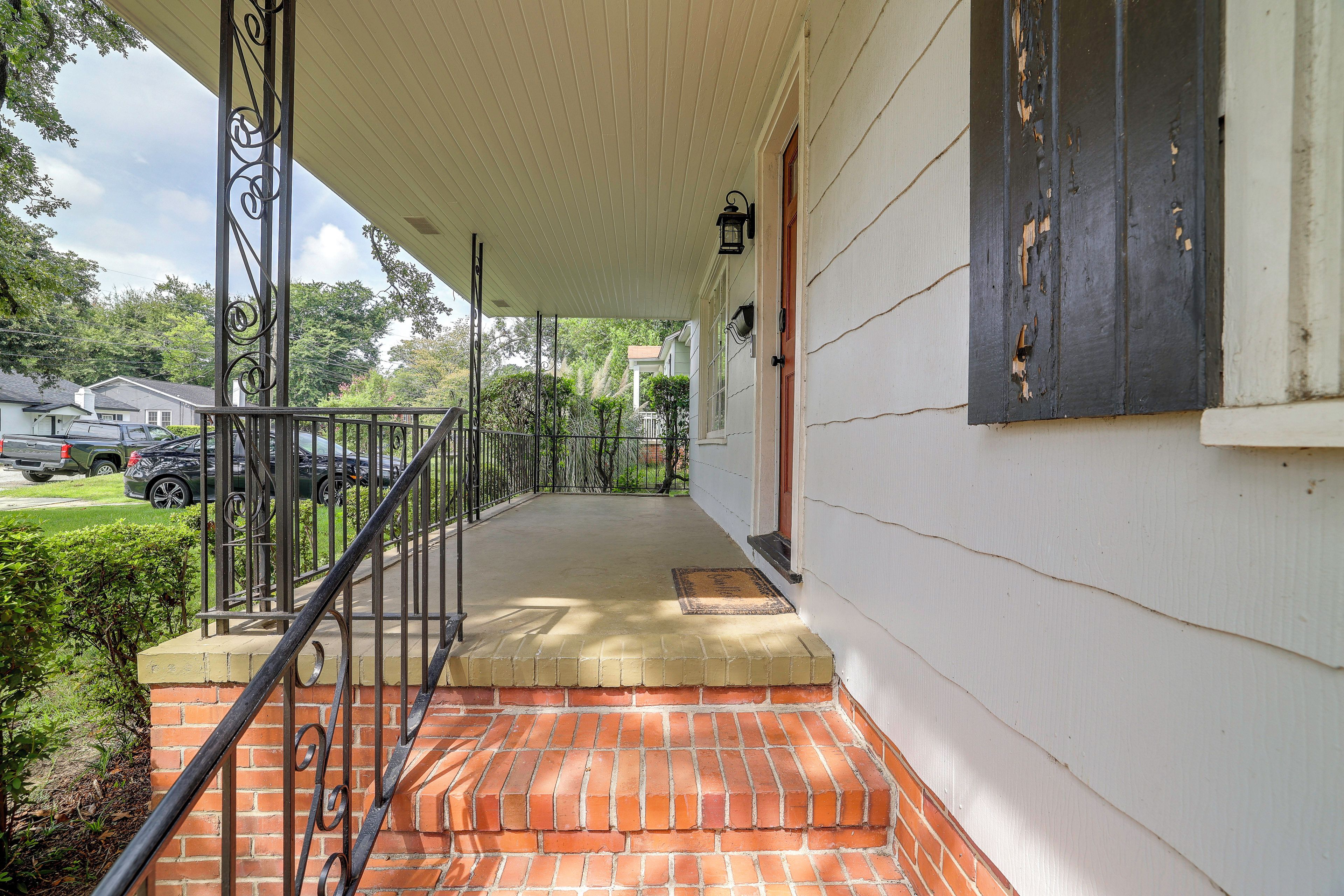A covered porch with a brick step leading to a door, a metal railing, and a view of a green lawn and parked cars.