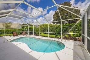 Ein Swimmingpool mit weißer Liegefläche, umgeben von einer überdachten Fläche mit Metallrahmen und Glasverkleidung, mit Blick auf eine Wohngegend mit Häusern und Grünflächen.