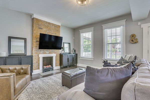 A living room with a brick wall, a flat-screen TV, a fireplace, and a large window with blinds.