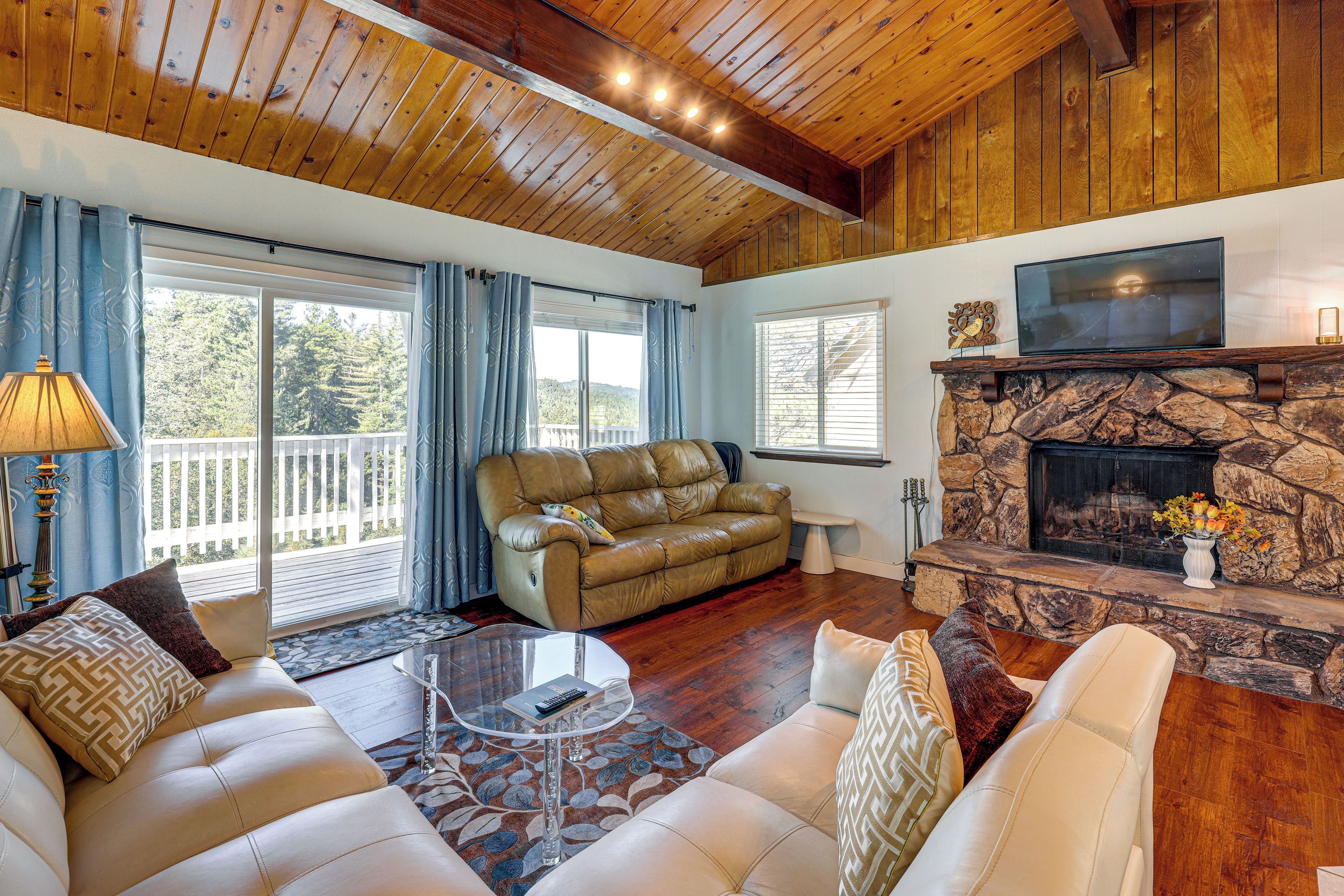 A cozy living room with a leather sofa, a television mounted above a stone fireplace, and a balcony with a view of trees.