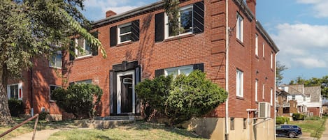 A two-story brick house with a front door, windows, and a small front yard with grass and shrubs.