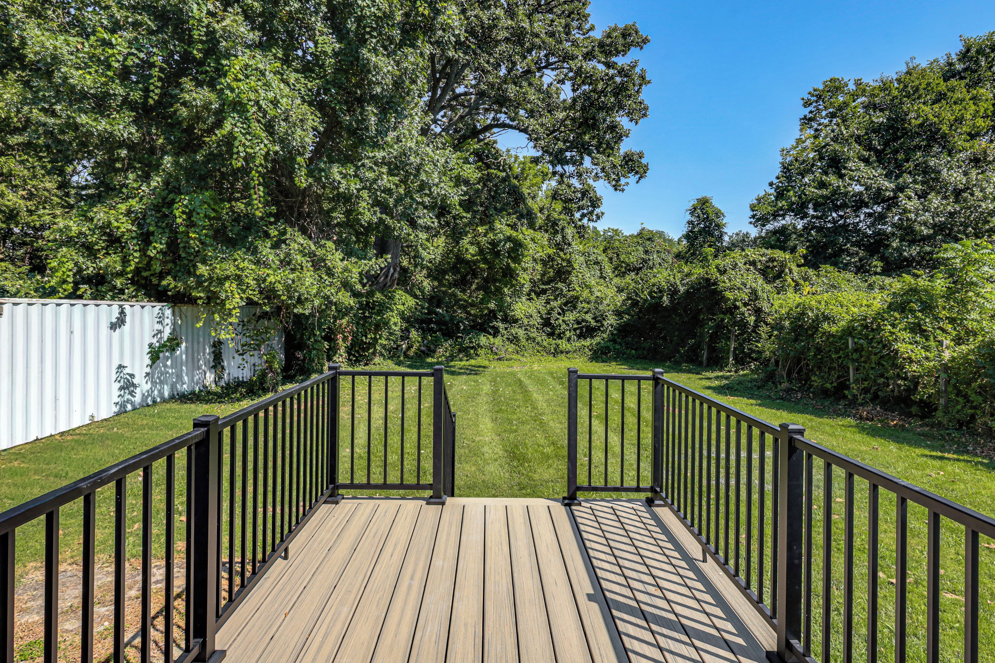 A wooden deck with black railings leading to a grassy area surrounded by trees.