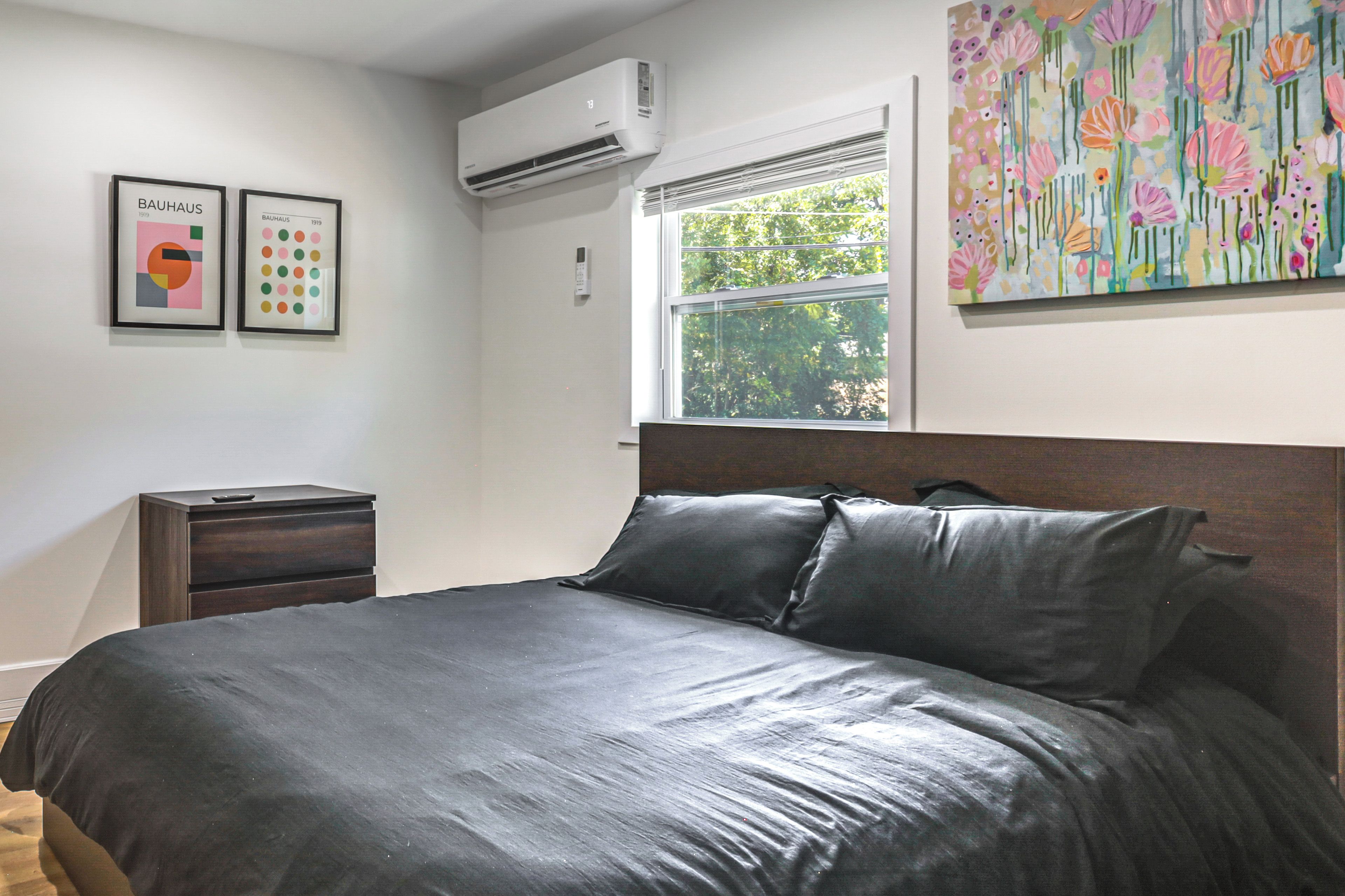 Ein Schlafzimmer mit einem großen Bett, einem Nachttisch, einer Klimaanlage, einem Fenster mit Jalousien und zwei gerahmten Kunstwerken an der Wand.