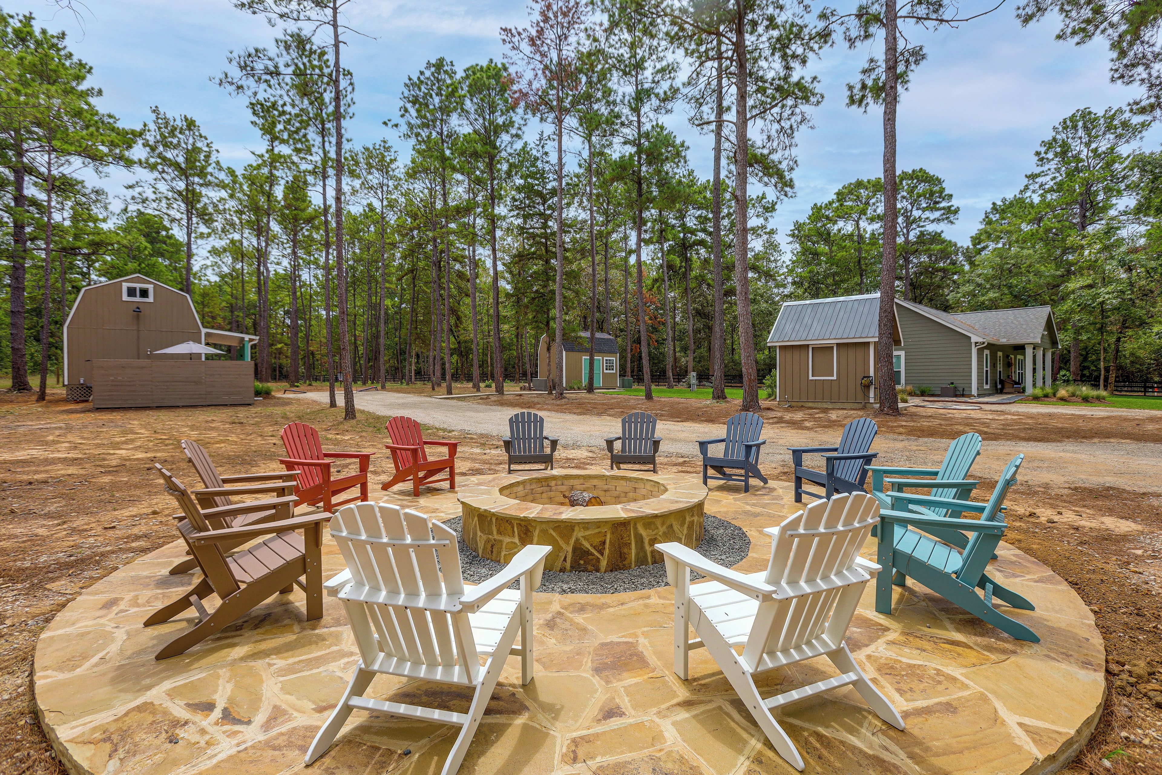 A stone fire pit surrounded by Adirondack chairs in a wooded outdoor area with a barn and a small cabin.