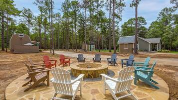 A stone fire pit surrounded by Adirondack chairs in a wooded outdoor area with a barn and a small cabin.