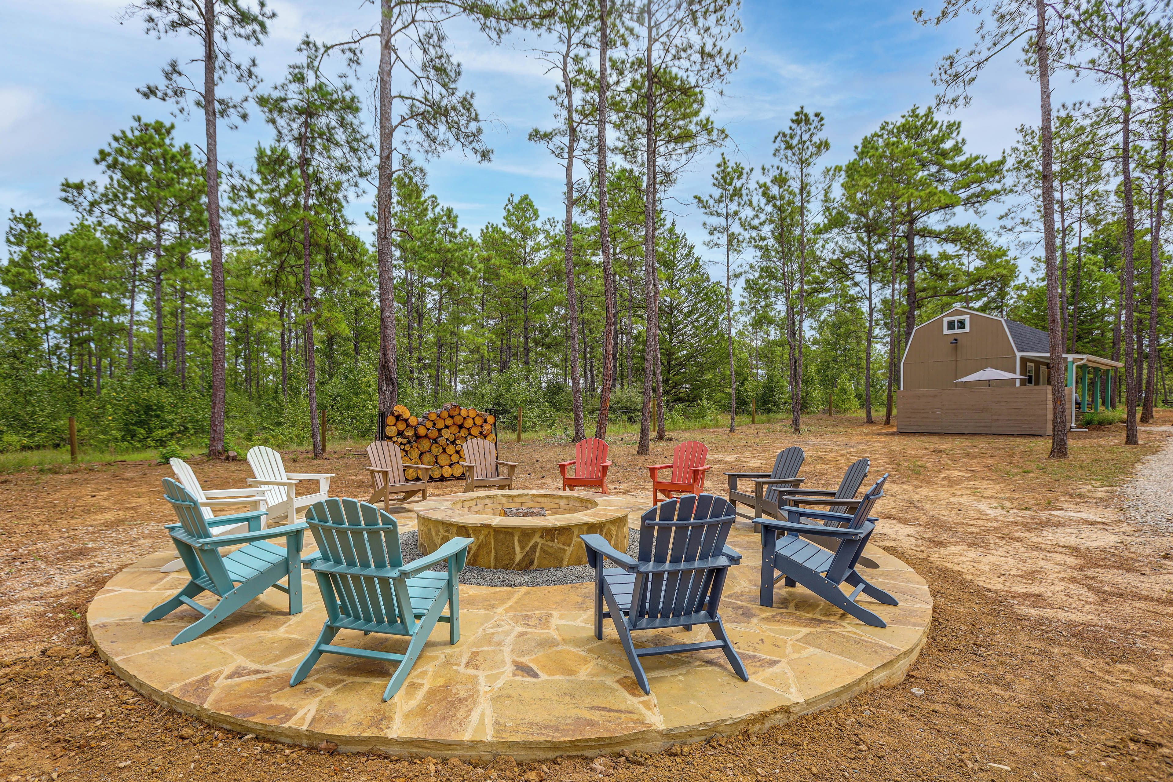 A circular stone fire pit surrounded by Adirondack chairs in a forest setting.