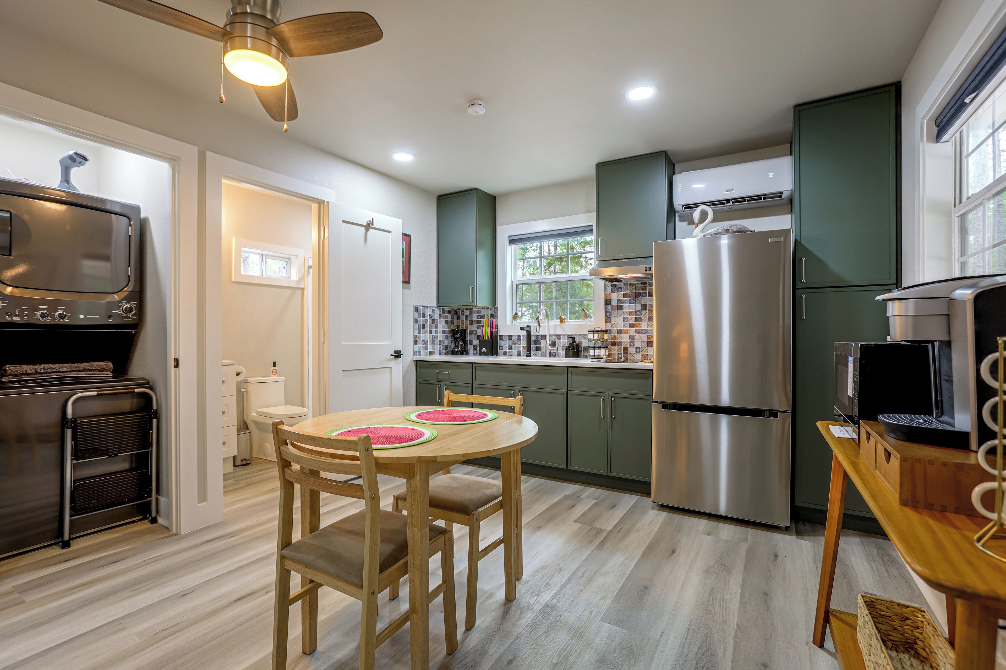A modern kitchen with a stainless steel refrigerator, a microwave, and a built-in oven. There is a dining table with chairs and a ceiling fan above.
