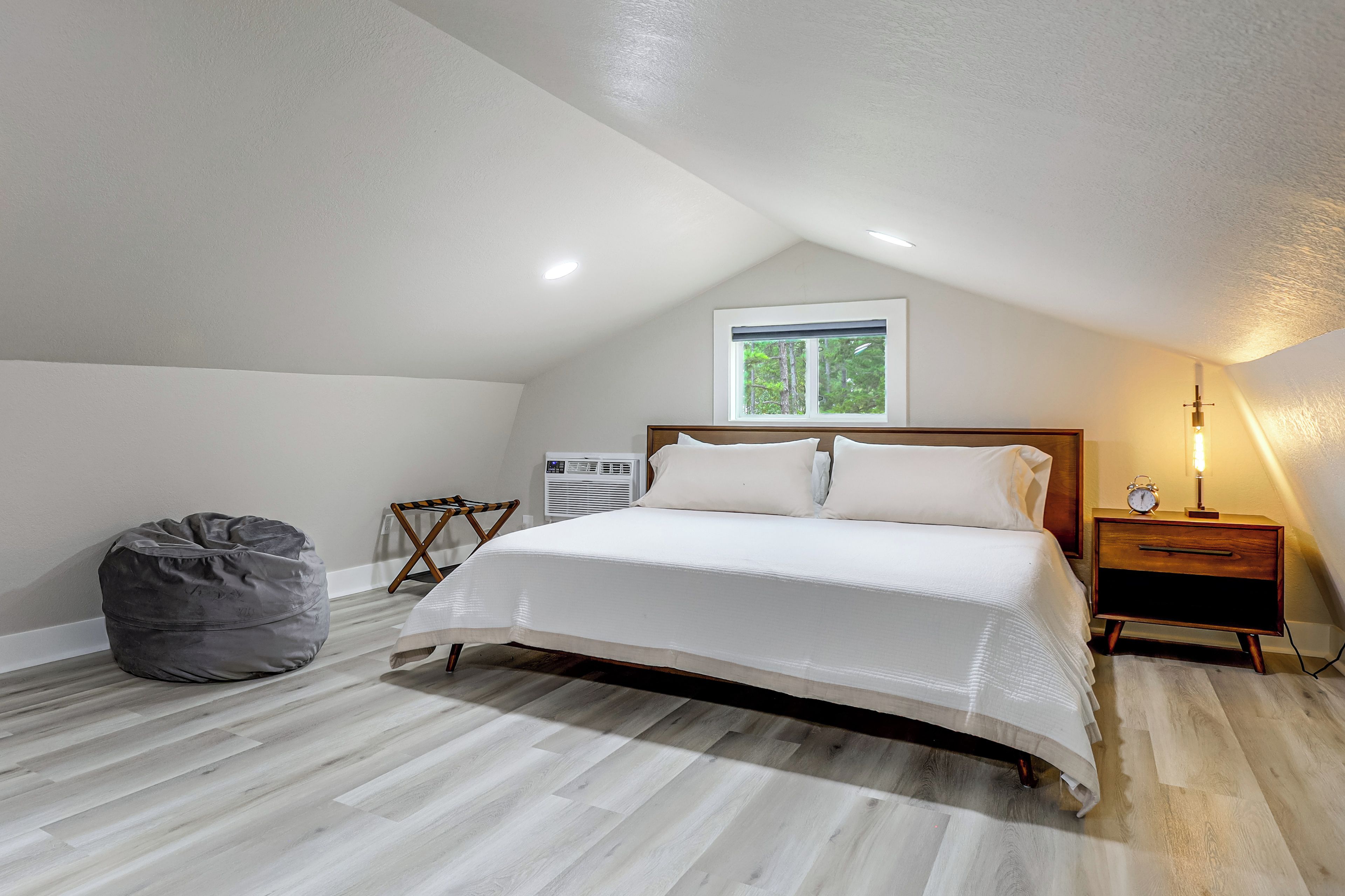 A bedroom with a bed, a bedside table with an alarm clock, a chair, a bean bag, and a window with a view of trees.