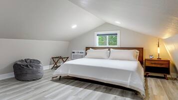 A bedroom with a bed, a bedside table with an alarm clock, a chair, a bean bag, and a window with a view of trees.