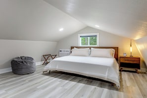 Un dormitorio con cama, una mesita de noche con un despertador, una silla, un sillón de peluche y una ventana con vistas a árboles.