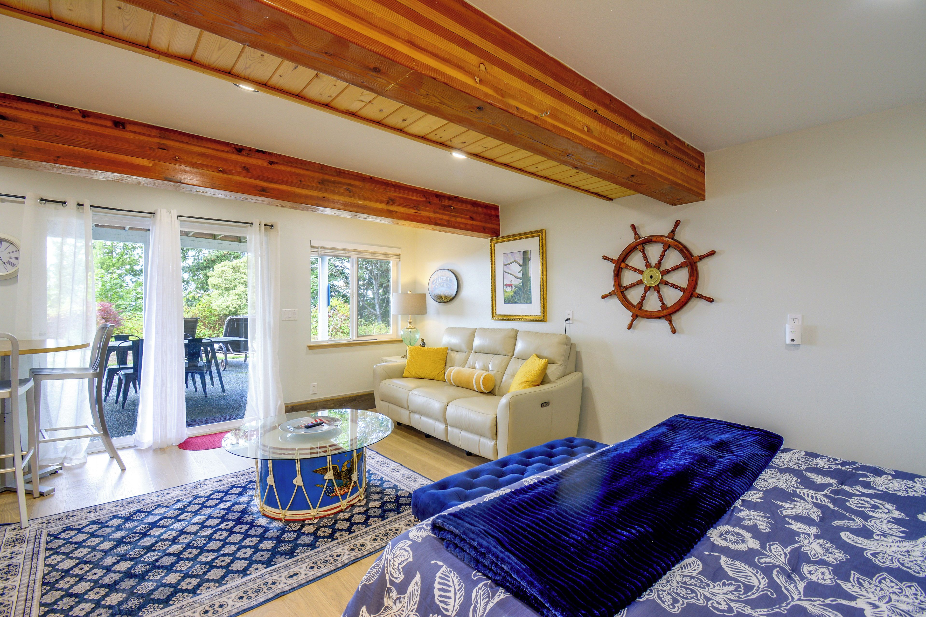 A living room with a wooden ceiling, a white sofa with yellow cushions, a blue patterned rug, and a ship's wheel decoration.