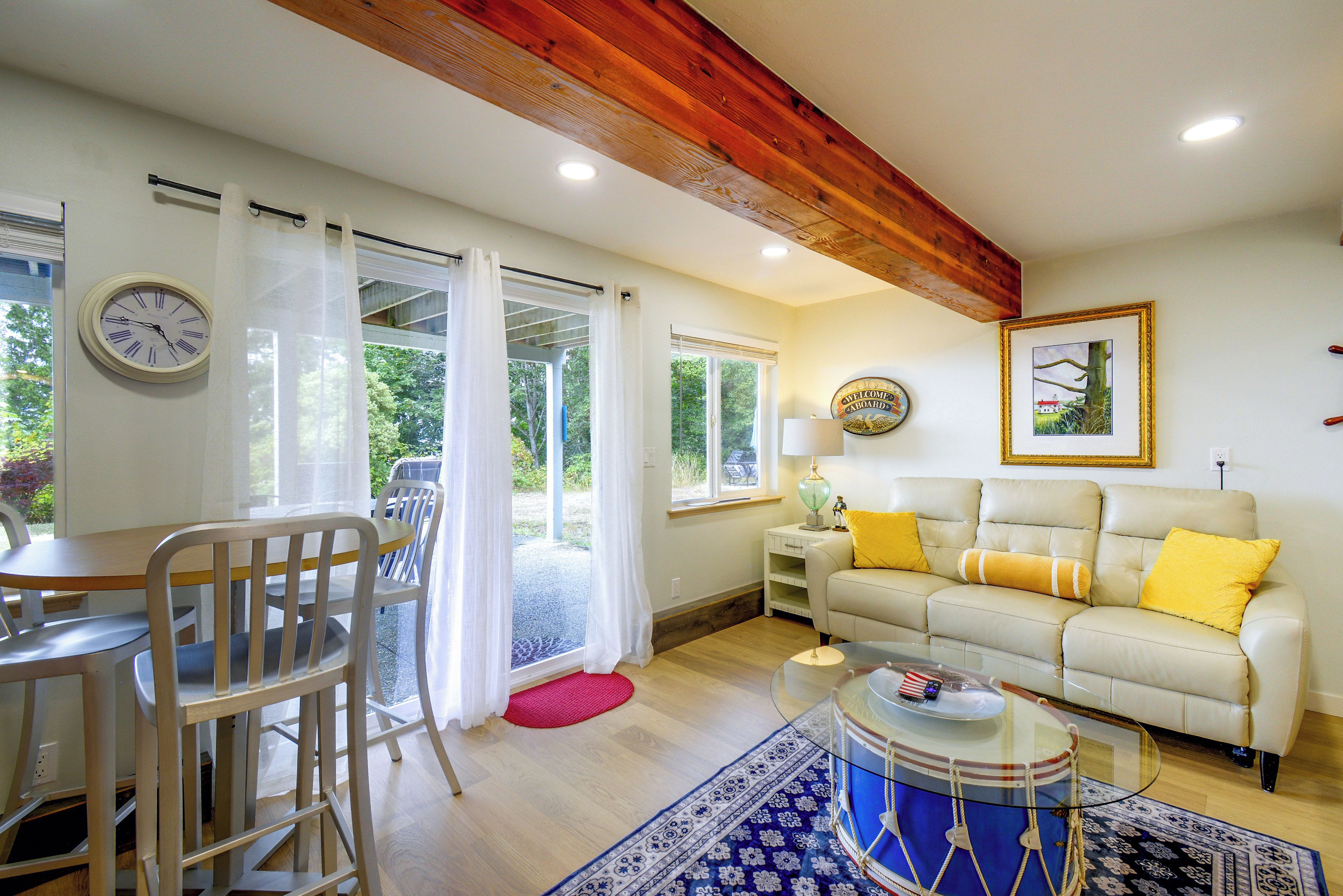 A room with a dining table, chairs, a sofa with yellow cushions, a drum, and a clock on the wall.