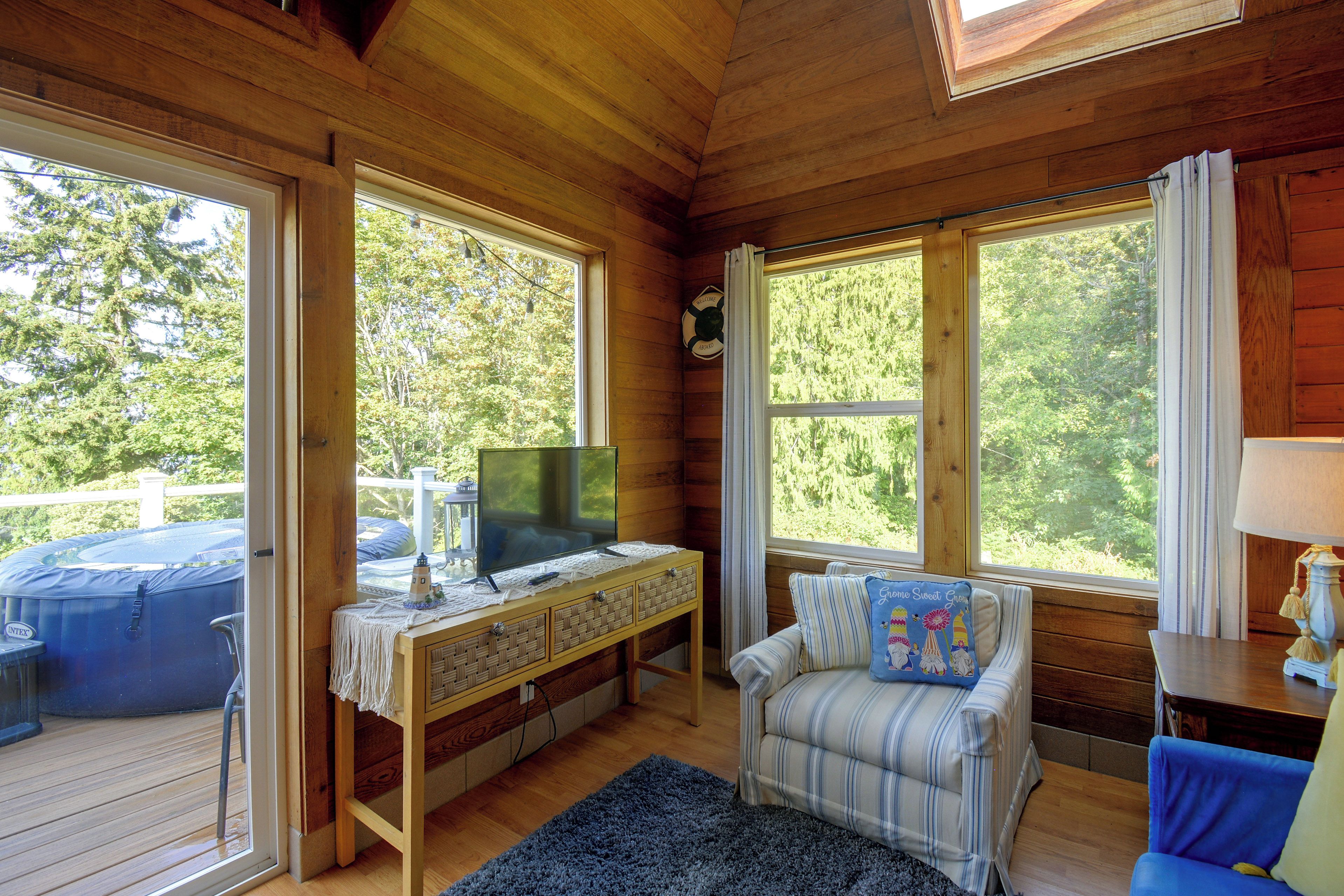 A wooden cabin interior with a television on a table, a sofa, and a window overlooking a deck.