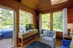 A wooden cabin interior with a television on a table, a sofa, and a window overlooking a deck. - Walk to Beach! Kitsap Coast Retreat w/ Hot Tub (Hansville)