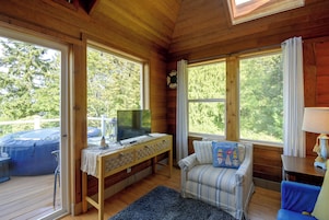 A wooden cabin interior with a television on a table, a sofa, and a window overlooking a deck.
