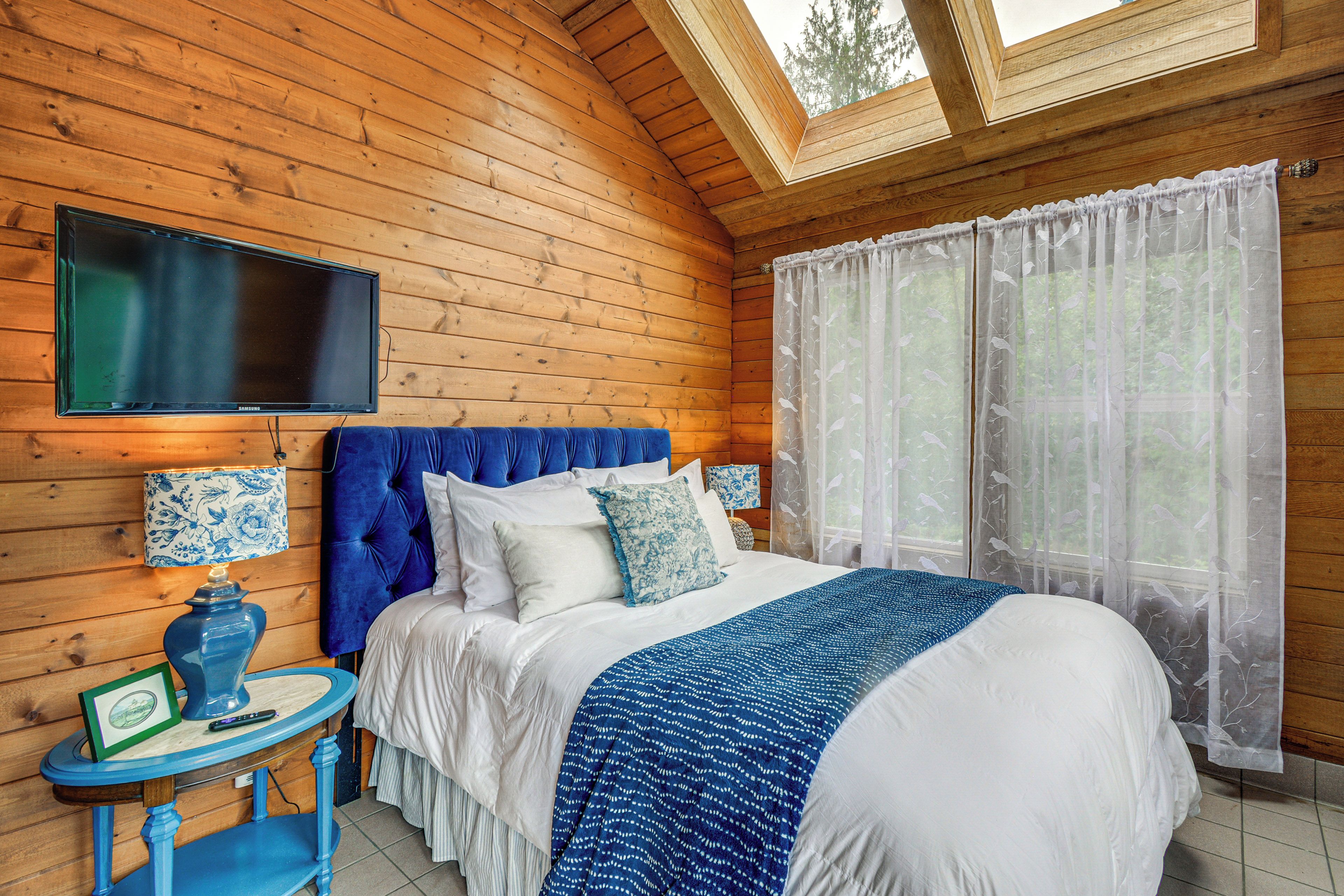 A bedroom with a bed, a television mounted on the wall, a window with sheer curtains, and a small table with a lamp.