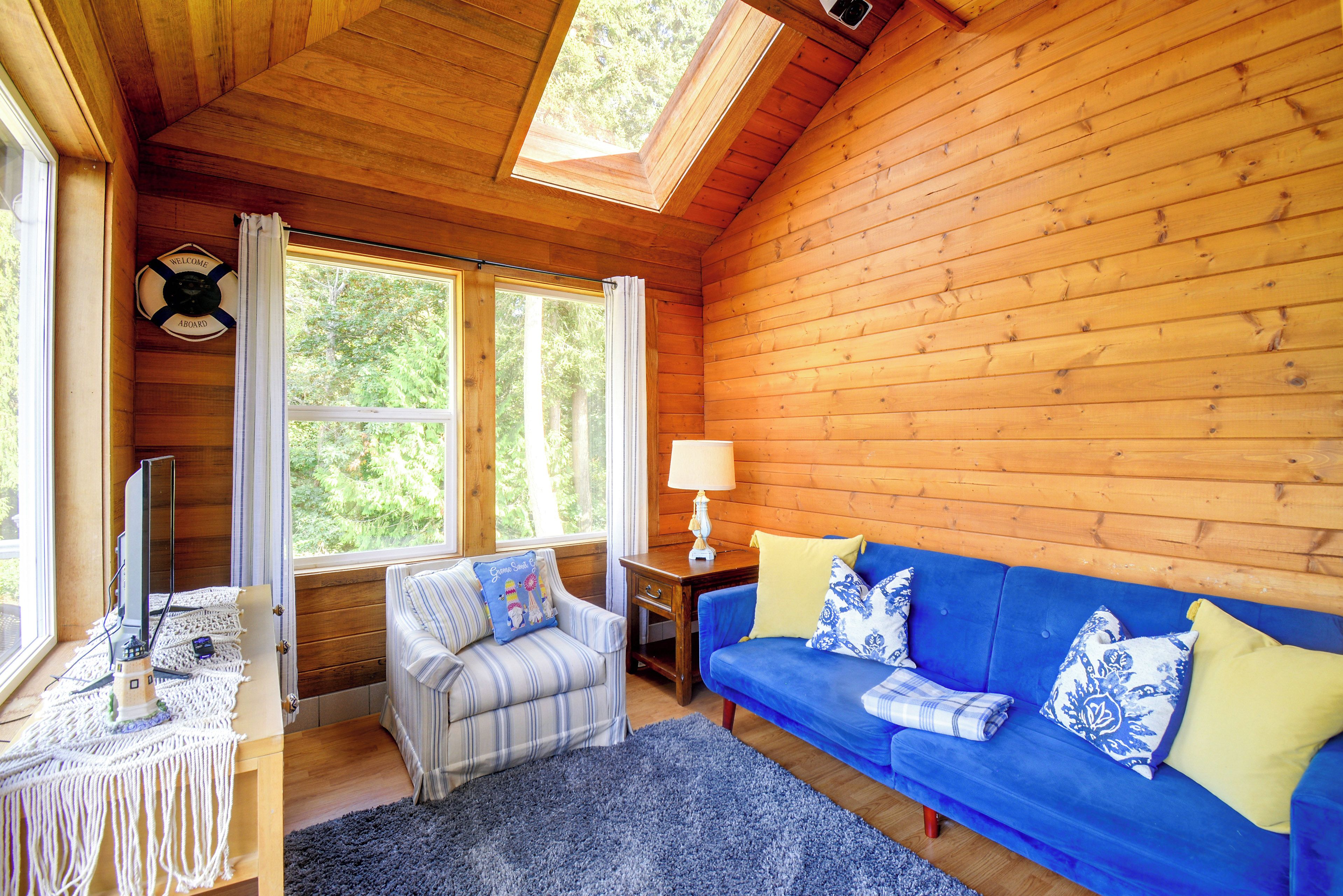 A cozy cabin interior with a blue sofa, a striped armchair, a small table with a lamp, and a television.