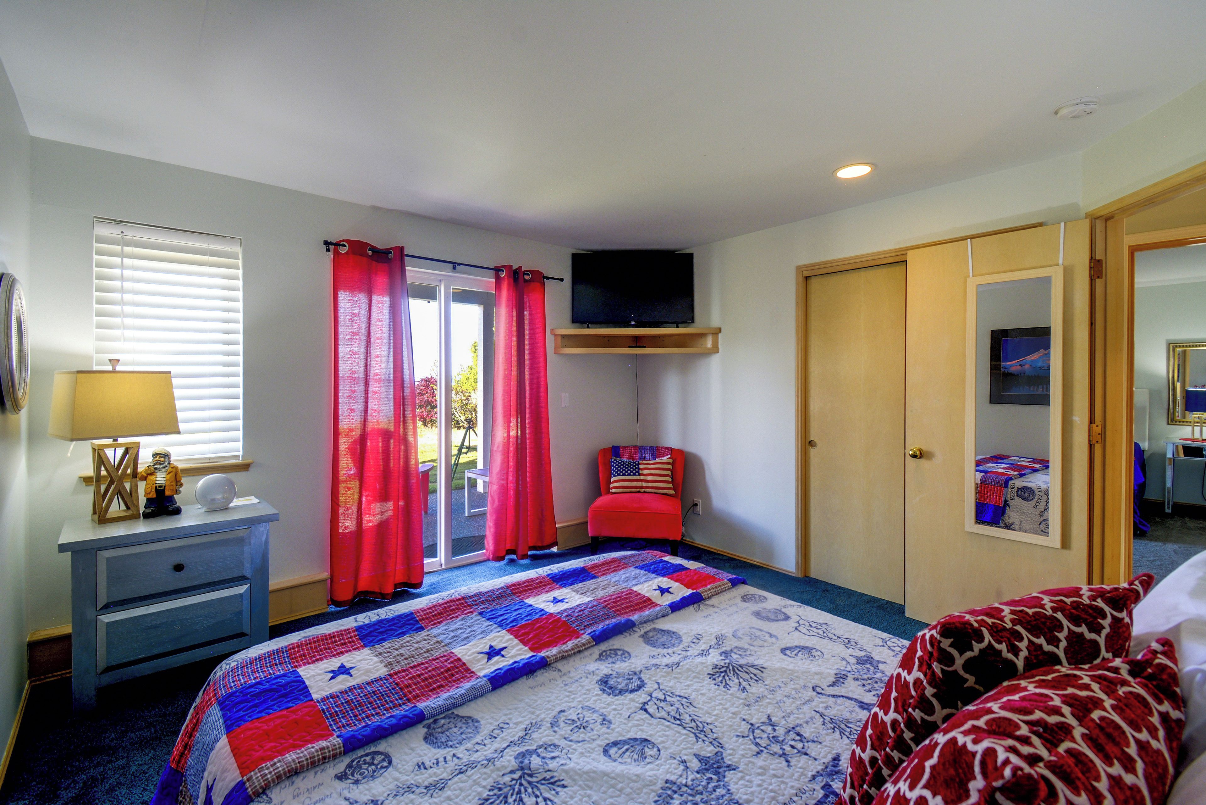 A bedroom with a bed, a television mounted on the wall, a desk with a lamp, and a window with blinds.